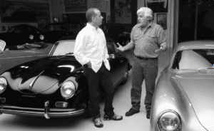 Two men standing and conversing in a garage surrounded by vintage cars, including a black and a silver Porsche. One man wears a white shirt and jeans, while the other sports a denim shirt and jeans.